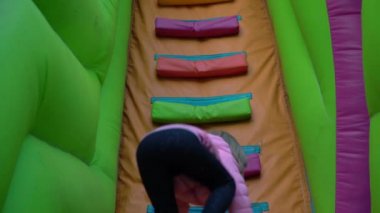 Bir kız çocuk parkında şişirilebilir bir kaydırağın üstünde merdivenlerden çıkıyor.