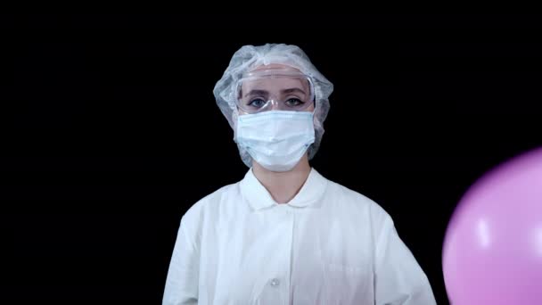 En sjuksköterska i skyddsutrustning håller en rosa ballong i handen och genomborrar den med en spruta — Stockvideo