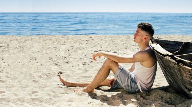 Beyaz T-shirt genç bir adam sahilde dinlenin. O eski teknenin oturuyor. O şapka elinde tutan ve uzak dreamly arıyorsunuz.