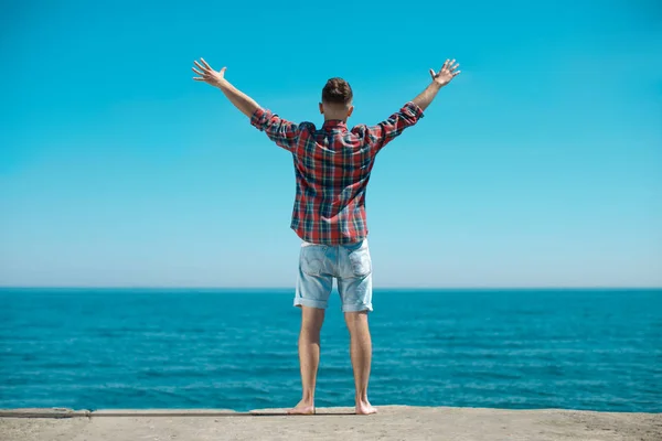 The guy in the checkered shirt is standing on the pier with his back to the camera. Before him there is a beautiful view of the spacious sea, he is happy and he lifts his arms upwards from delight.