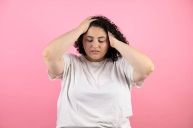 Genç ve güzel bir kadın yalnız pembe arka planda duruyor. Baş ağrısı, çaresiz ve stresli. Çünkü elleri başının üstünde acı ve migren var.