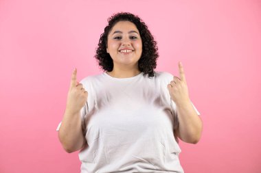 Soyutlanmış pembe arka planda güzel bir kadın. Gülümsüyor, kameraya bakıyor, parmaklarıyla işaret ediyor ve kollarını kaldırıyor.