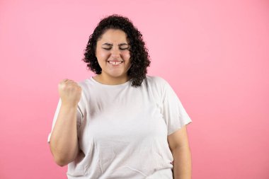 Genç ve güzel bir kadın pembe arka planda çok mutlu ve heyecanlı. Kolunu kaldırmış ve dişlerini sıkarak kazanan bir jest yapıyor..