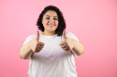 Genç ve güzel bir kadın pembe arka planda durmuş gülümsüyor ve baş parmaklarıyla işaret veriyor.