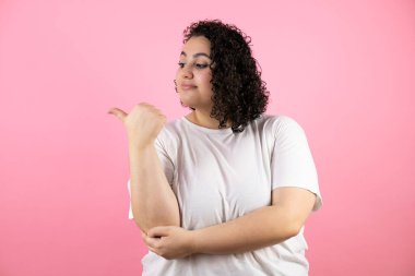 Soyutlanmış pembe arka planda genç ve güzel bir kadın başparmağını yana uzatıyor mutlu bir şekilde gülümsüyor.