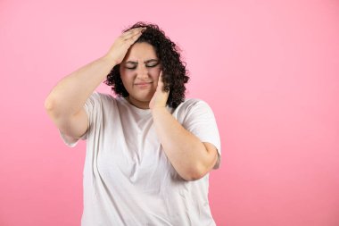Genç ve güzel bir kadın yalnız pembe arka planda duruyor. Baş ağrısı, çaresiz ve stresli. Çünkü elleri başının üstünde acı ve migren var.