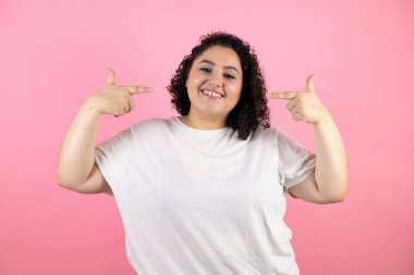 Soyutlanmış pembe arka planda genç ve güzel bir kadın gülümsüyor ve parmaklarıyla ve ağzıyla gösterip işaret ediyor.
