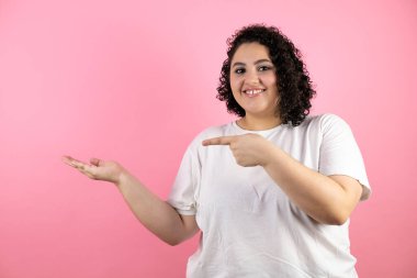 Genç, güzel bir kadın pembe arka planda duruyor. Gülümsüyor ve elindeki bir şeyi gösteriyor.