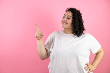 İzole edilmiş pembe arka planda duran genç ve güzel bir kadın gülümseyen ve yan bakan eli ve parmağıyla kenarı işaret ediyor..