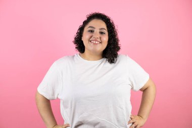 Genç, güzel bir kadın pembe arka planda duruyor. Kolları belinde gülümsüyor.