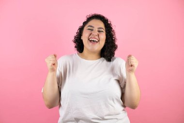 Genç ve güzel bir kadın pembe arka planda çok mutlu ve heyecanlı kollarını kaldırmış, gülümseyen ve başarı için çığlık atan kazanan bir jest yapıyor..