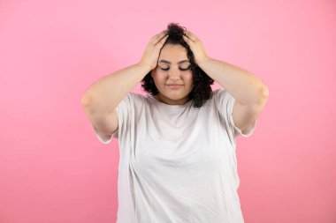 Genç ve güzel bir kadın yalnız pembe arka planda duruyor. Baş ağrısı, çaresiz ve stresli. Çünkü elleri başının üstünde acı ve migren var.