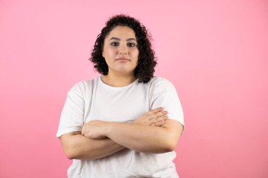 İzole edilmiş pembe arka planda duran genç ve güzel bir kadın kollarını kavuşturup kameraya çok ciddi bir şekilde bakıyor.