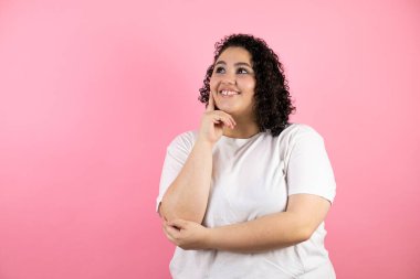 Genç, güzel bir kadın pembe arka planda duruyor. Kendine güvenen, kollarını kavuşturmuş ve çenesini kaldırmış gülümseyen bir kadın.