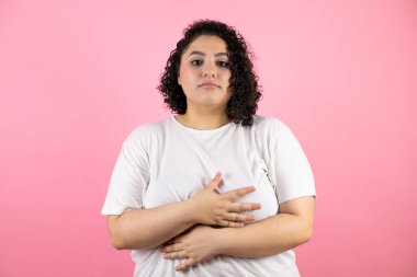 Genç ve güzel bir kadın karnına dokunuyor ve önüne bakıyor. Pembe arka planda günlük beyaz tişört giyiyor.