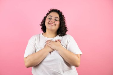 Pembe arka planda duran genç ve güzel bir kadın elleri göğsünde, gözleri kapalı ve yüzünde minnet dolu bir jest ile gülümsüyor..