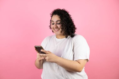 Genç ve güzel bir kadın telefonuyla mesaj atıyor. Pembe arka planda gülümsüyor..