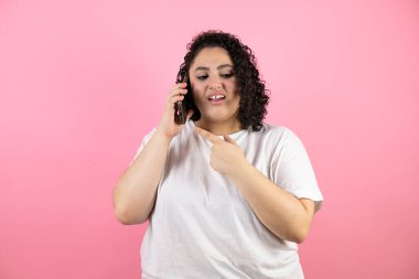 Genç ve güzel bir kadın telefonda konuşuyor. Kafası karışık bir ifadeyle ve diğer eliyle pembe arka planda onu işaret ederek..