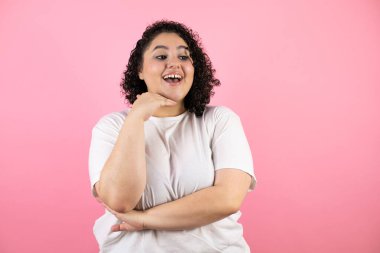 Genç, güzel bir kadın pembe arka planda duruyor. Kendine güvenen, kollarını kavuşturmuş ve çenesini kaldırmış gülümseyen bir kadın.