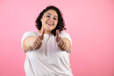 Genç ve güzel bir kadın pembe arka planda durmuş gülümsüyor ve baş parmaklarıyla işaret veriyor.