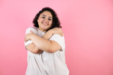 Soyutlanmış pembe arka planda duran genç ve güzel bir kadın. Kendine sarılıyor. Mutlu ve pozitif.