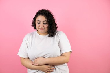 Genç ve güzel bir kadın karnına dokunuyor ve önüne bakıyor. Mavi arka planda günlük beyaz tişört giyiyor.