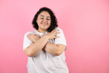 Soyutlanmış pembe arka planda duran genç ve güzel bir kadın. Kendine sarılıyor. Mutlu ve pozitif.