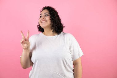 İzole edilmiş pembe arka planda gülümseyen ve zafer işareti yapan genç esmer kadın. İki numara..