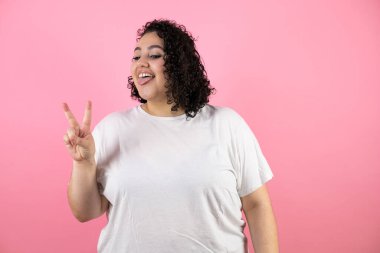 İzole edilmiş pembe arka planda gülümseyen ve zafer işareti yapan genç esmer kadın. İki numara..