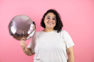 Genç ve güzel bir kadın pembe arka planda durup gülümsüyor ve elinde bir balon tutuyor.