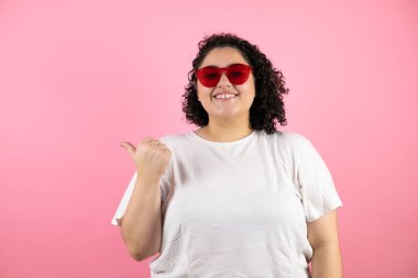 Güneş gözlüğü takan genç ve güzel bir kadın pembe arka planda duruyor. Gülümsüyor ve başparmağıyla işaret ediyor.