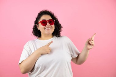 Güneş gözlüğü takan genç ve güzel bir kadın pembe arka planda duruyor. Gülümsüyor, elleri ve parmakları yana doğru uzanıyor.