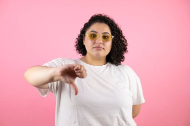 İzole edilmiş pembe arka planda güneş gözlüğü takan genç ve güzel bir kadın. Kızgın bir yüzü var.