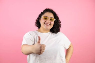 Güneş gözlüğü takan genç ve güzel bir kadın pembe arka planda duruyor gülümsüyor ve baş parmağıyla işaret veriyor.