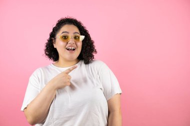 Güneş gözlüğü takan genç ve güzel bir kadın pembe arka planda duruyor. Gülümsüyor, şaşırıyor ve elleri ve parmaklarıyla işaret ediyor.