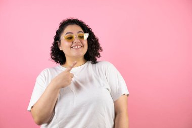 Güneş gözlüğü takan genç ve güzel bir kadın pembe arka planda durup gülümsüyor.