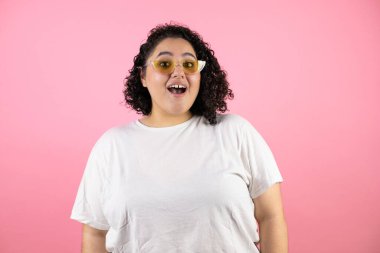 Güneş gözlüğü takan genç ve güzel bir kadın soyutlanmış pembe arka planda duruyor.