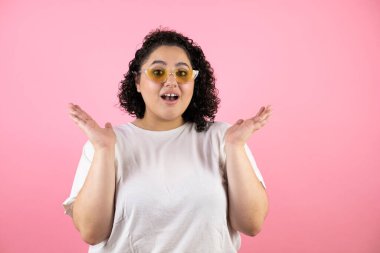 Güneş gözlüğü takan genç ve güzel bir kadın soyutlanmış pembe arka planda duruyor.