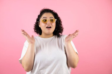 Güneş gözlüğü takan genç ve güzel bir kadın soyutlanmış pembe arka planda duruyor.