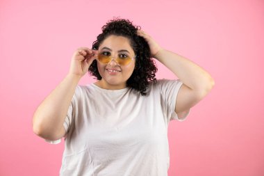 İzole edilmiş pembe arka planda güneş gözlüğü takan güzel bir kadın gülümsüyor, alıyor ve güneş gözlüğünü indiriyor.