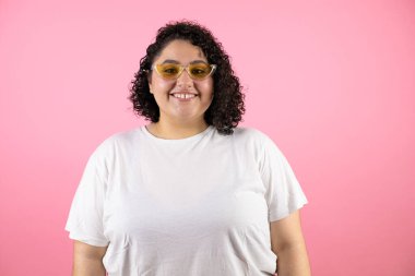 Güneş gözlüğü takan genç ve güzel bir kadın soyutlanmış pembe arka planda gülümsüyor.