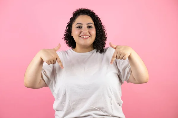 若いです美しいです女性上の隔離されたピンクの背景笑顔 カメラを見て 指で下を指して腕を上げました — ストック写真