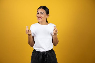 Zararlı bir suratı olan genç ve güzel bir kadın sarı arka planda parmaklarıyla para işareti yapıyor. Beyaz gömlek ve siyah şort giyip ön tarafa bakıyordu..