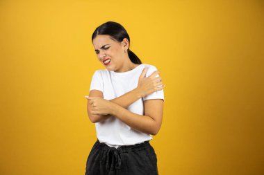 Omzunda ağrı ve sarı arka planda acı dolu bir ifade olan güzel bir kadın. Beyaz tişört ve siyah şort giyiyor..