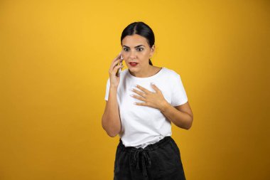 Sarı arka planda endişeli bir ifadeyle telefonda konuşan genç ve güzel bir kadın. Beyaz tişört, siyah şort ve kırmızı ruj giyiyor.