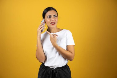 Kafası karışık bir ifadeyle telefonda konuşan genç ve güzel bir kadın diğer eliyle sarı arka planda ona işaret ediyor. Beyaz gömlek, siyah şort ve kırmızı ruj giyiyor.