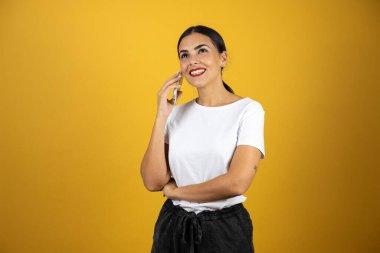 Kafası karışık bir ifadeyle telefonda konuşan genç ve güzel bir kadın diğer eliyle sarı arka planda ona işaret ediyor. Beyaz gömlek, siyah şort ve kırmızı ruj giyiyor.