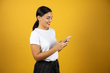 Genç, güzel bir kadın, sarı arka planda gülümserken çok mutlu bir şekilde telefonuyla mesajlaşıyor. Beyaz tişört, siyah şort ve kırmızı ruj giyiyor.