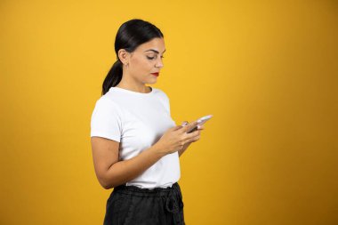 Genç ve güzel bir kadın sarı arka planda telefonuyla mesajlaşıyor. Beyaz gömlek, siyah şort ve kırmızı ruj giyiyor.