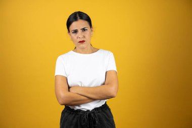 İzole edilmiş sarı bir arka planda duran genç ve güzel bir kadın kollarını kavuşturup ön tarafa çok kızgın bakıyor. Endişeli ifade